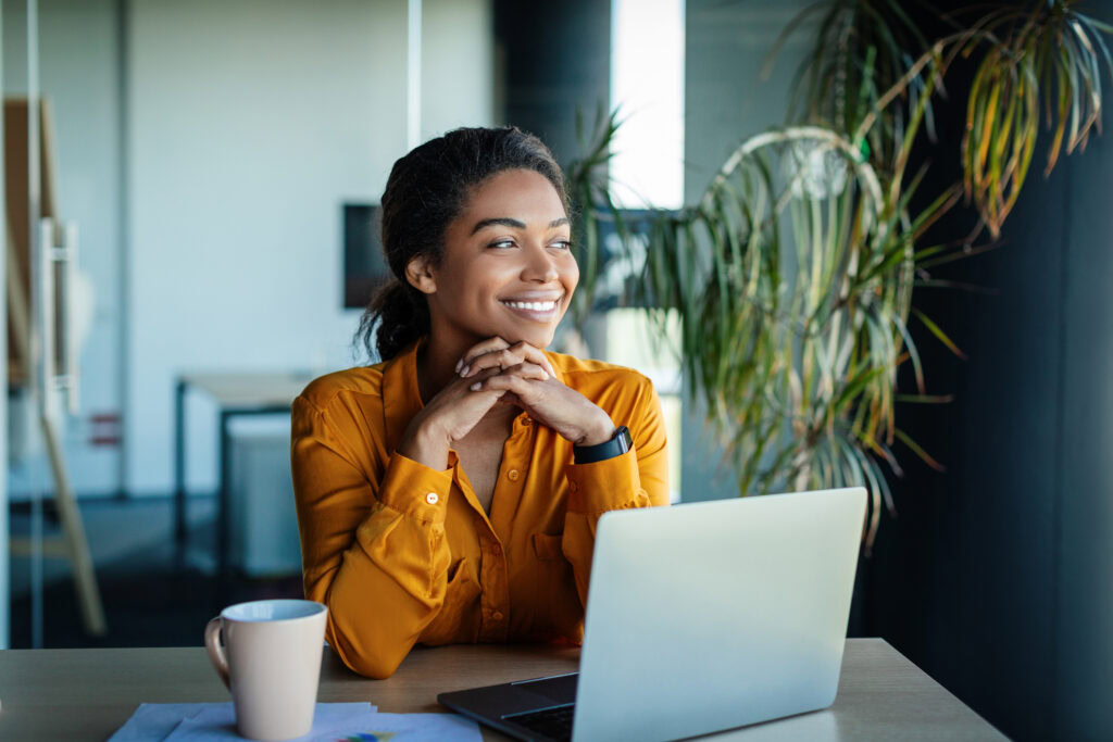 Portrait Of Dreamy African American Businesswoman Sitting At Desk In Office And Thinking While Working On Laptop